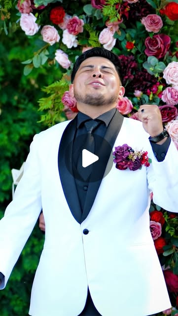 Groom in a white tuxedo in front of a wall of roses